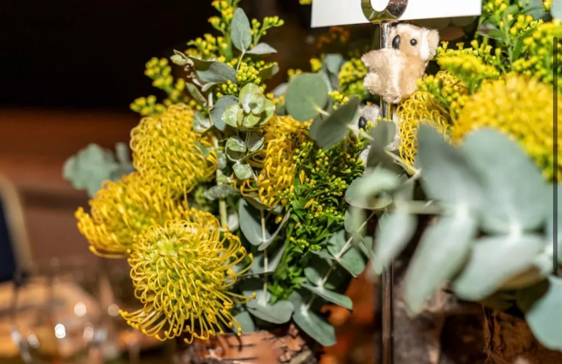 A centrepiece of yellow pincushion proteas, wattle and eucalyptus in a birch-bark vase, a small koala clipped to the table card above it