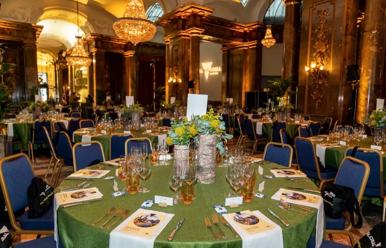 The hall before dinner, round tables laid under chandeliers between gilded columns, with a lit screen at the far end