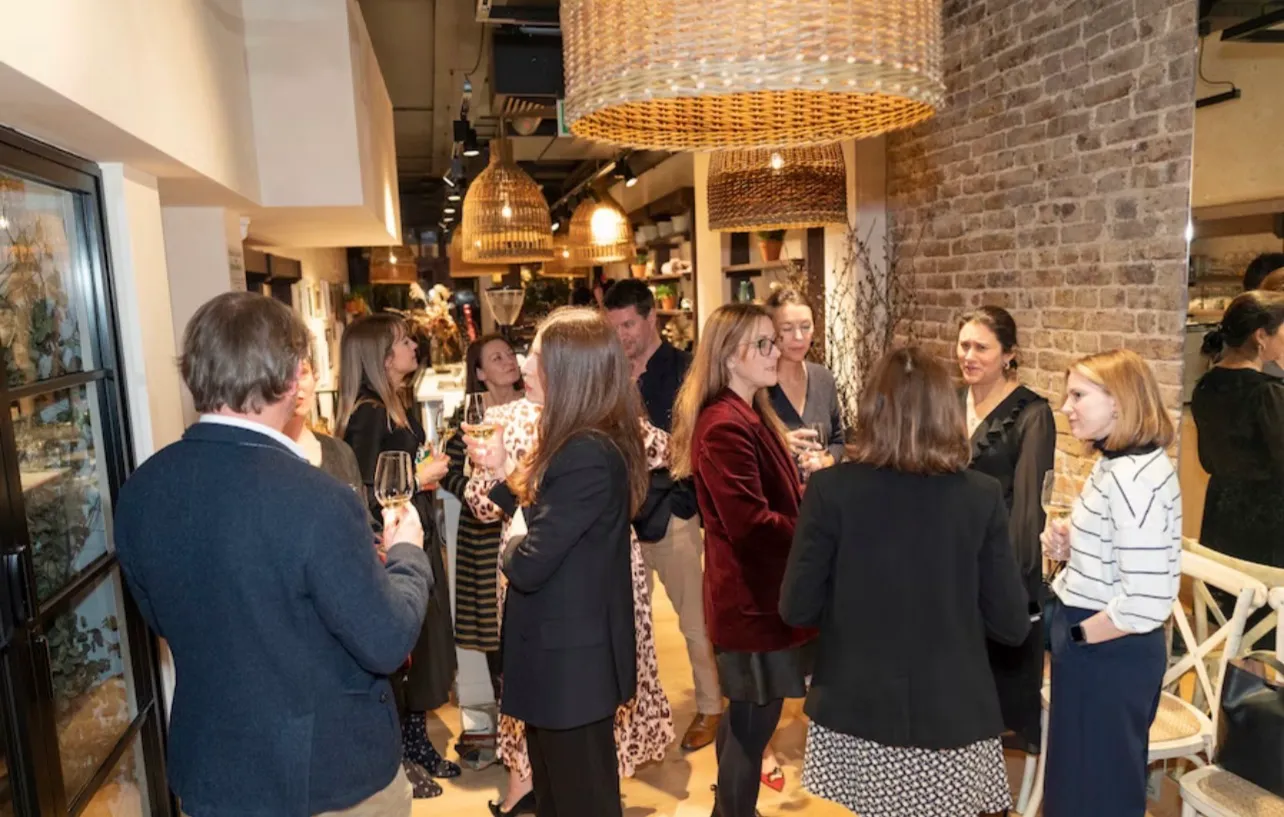 Guests standing with drinks in a shop showroom, talking in groups under woven pendant lights against exposed brick