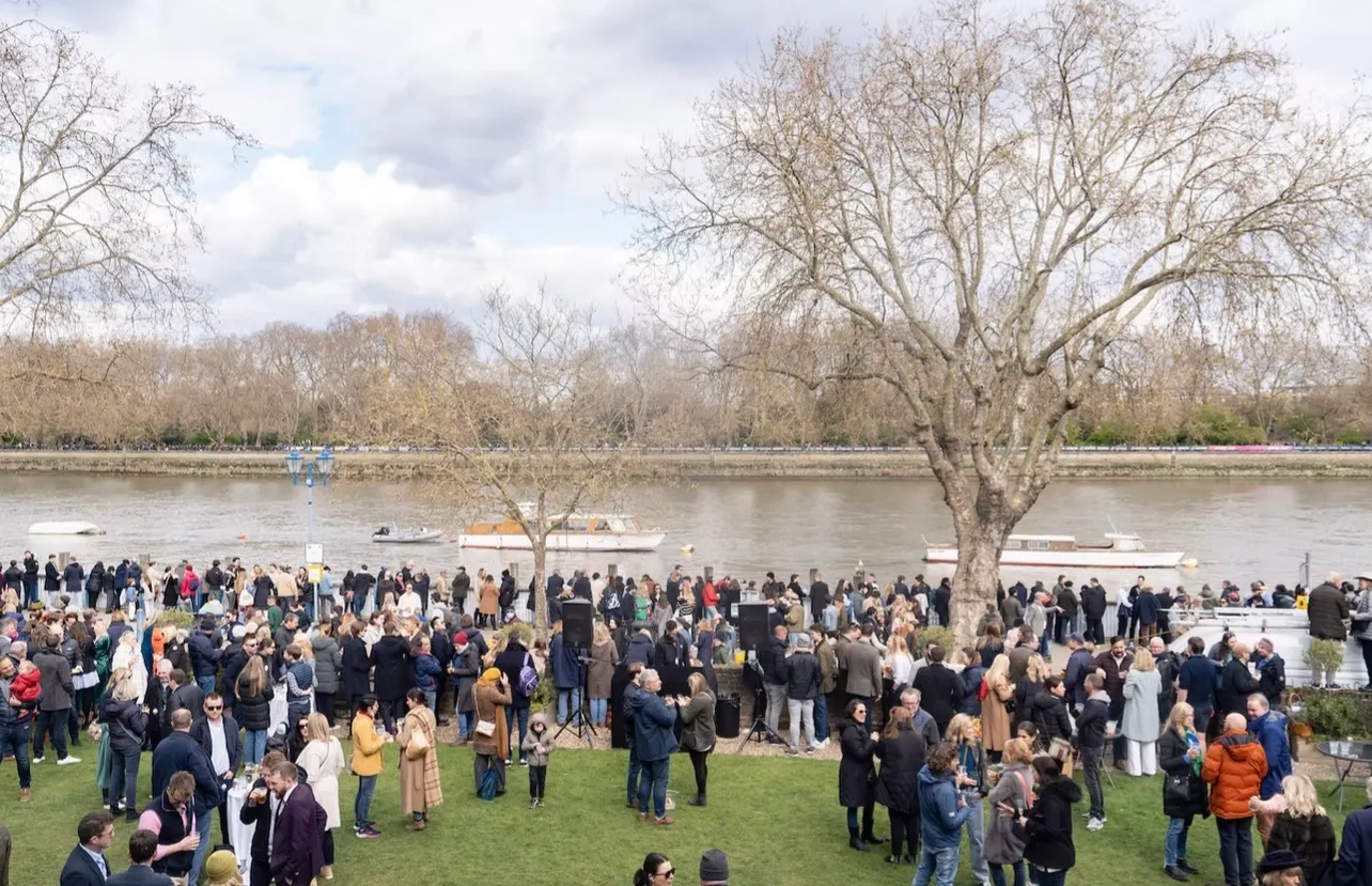 A crowd filling the lawn and the river wall on Boat Race day, drinks in hand and launches out on the Thames beyond