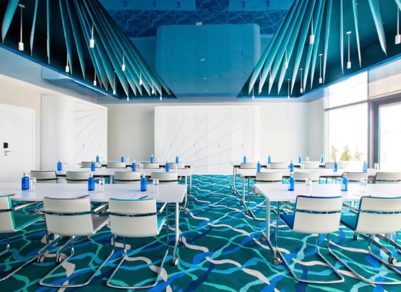 A meeting room set in rows for a day session, water and glasses laid at every place, under a slatted blue ceiling and full-height windows