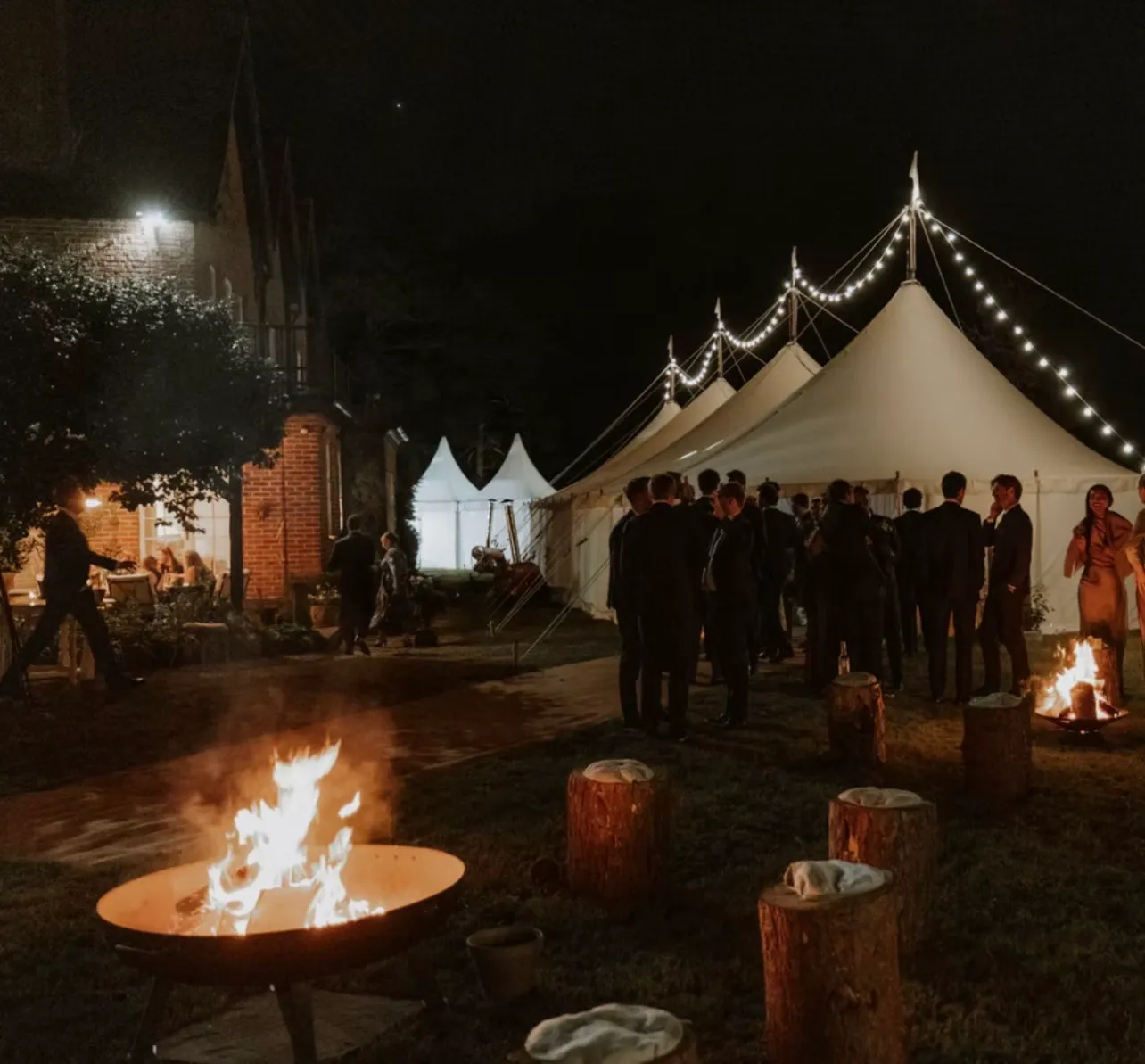 The marquee lit from within after dark with guests gathered at its open side, fire pits burning on the lawn and log seats set around them