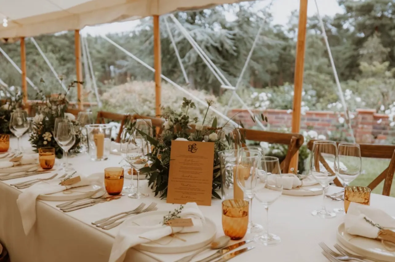 A place setting down one side of a trestle table, a printed menu standing against foliage and amber glasses, the walled garden beyond the marquee