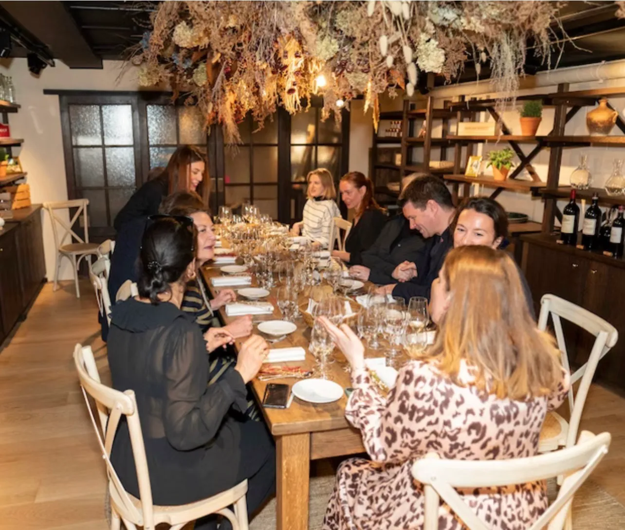 Guests seated along both sides of the long table in conversation, glasses poured in front of them and the dried flowers overhead
