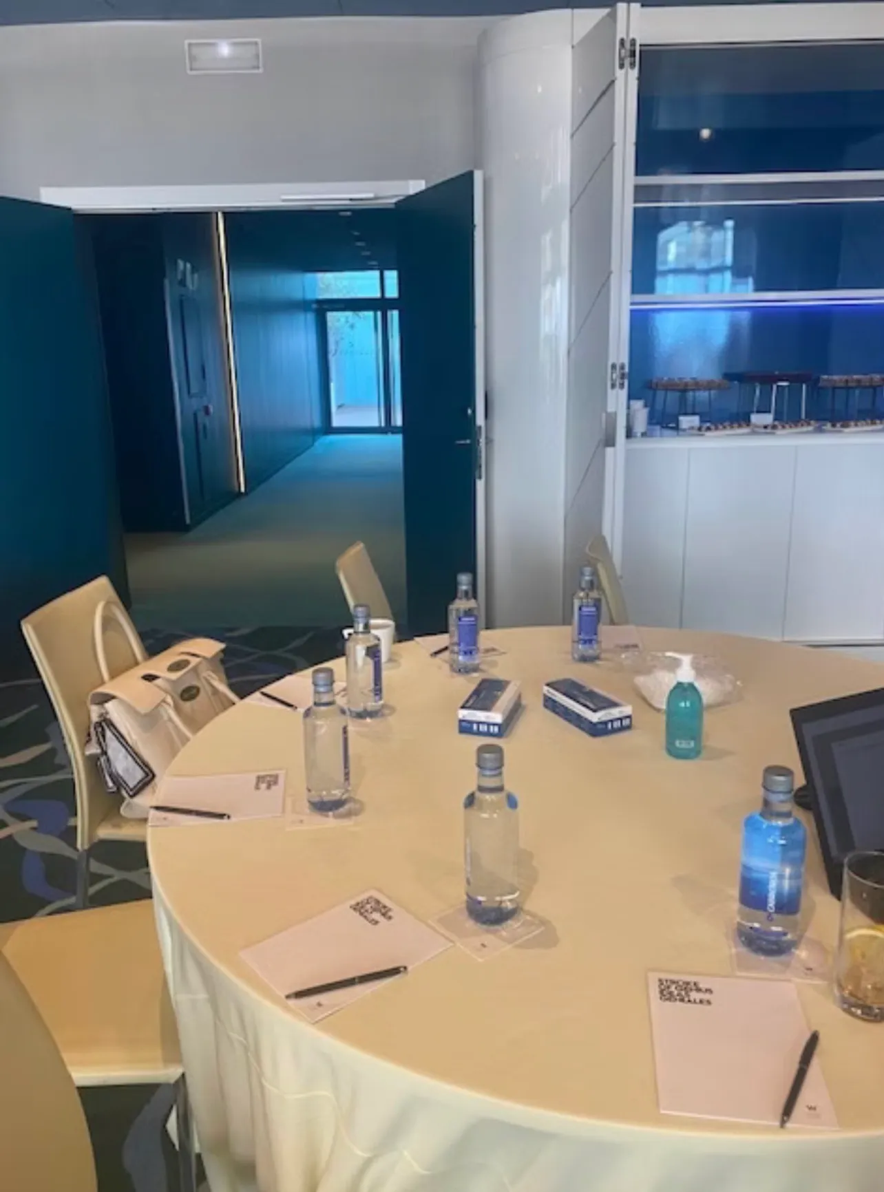 A round table laid for a small group with notepads, pens and bottled water at each place, a door standing open onto the corridor behind