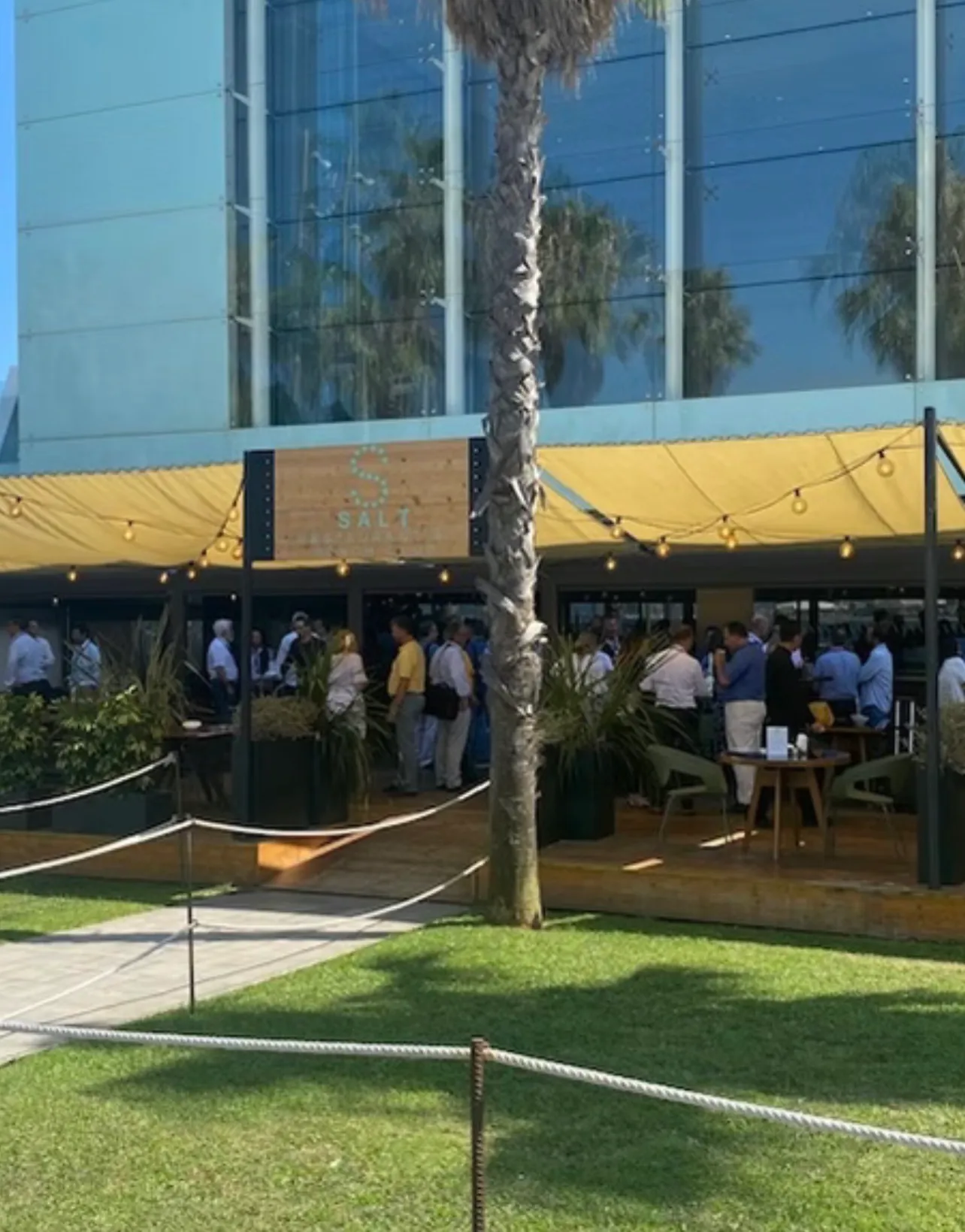 Delegates standing and talking in groups on a decked terrace under a yellow awning and festoon lights, a palm in the foreground