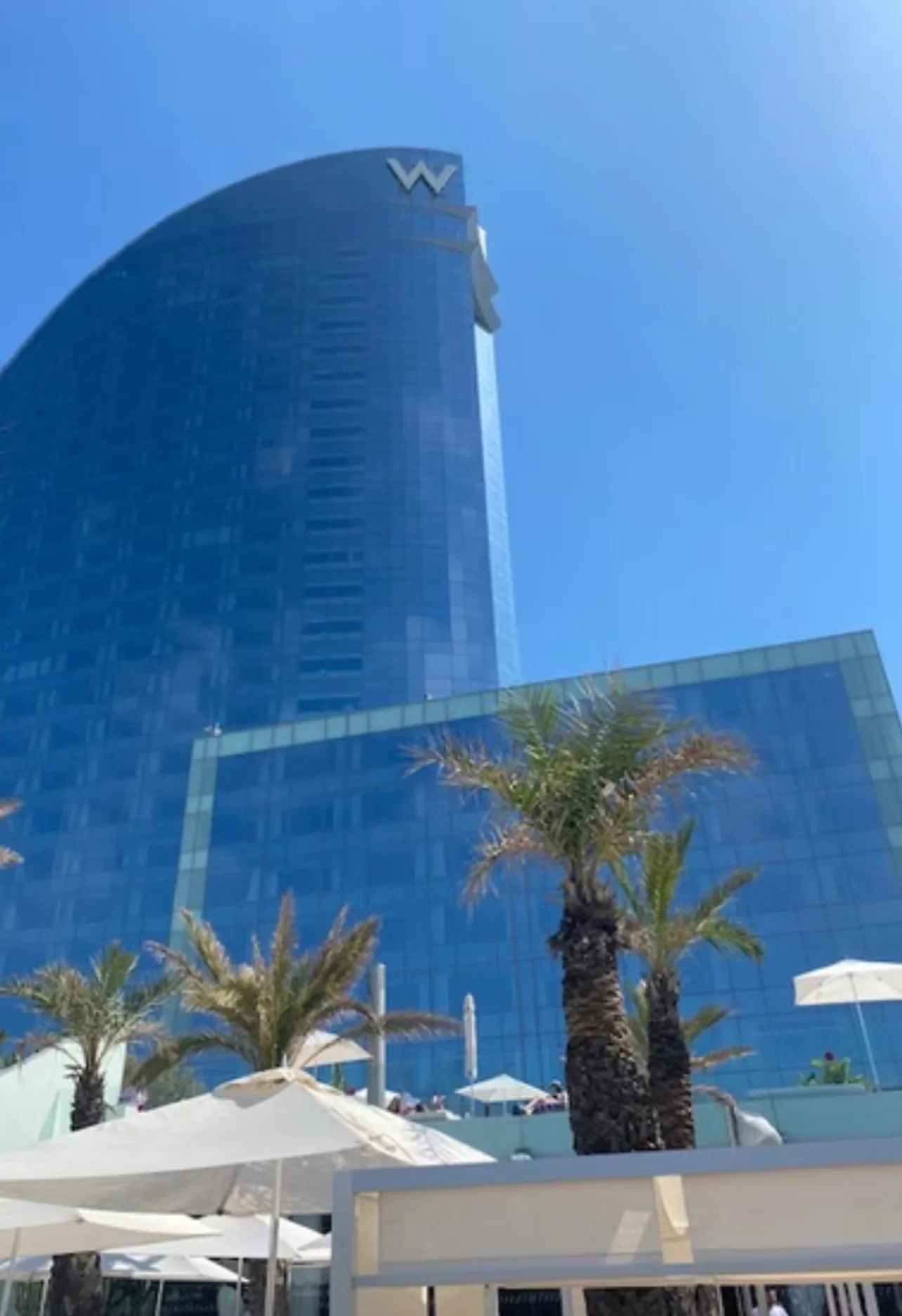 The tower seen from below against a clear sky, its W sign at the top and palms and white parasols on the deck in front of it